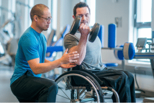 Homem cadeirante realizando exercício com halteres sob supervisão de um profissional em academia.